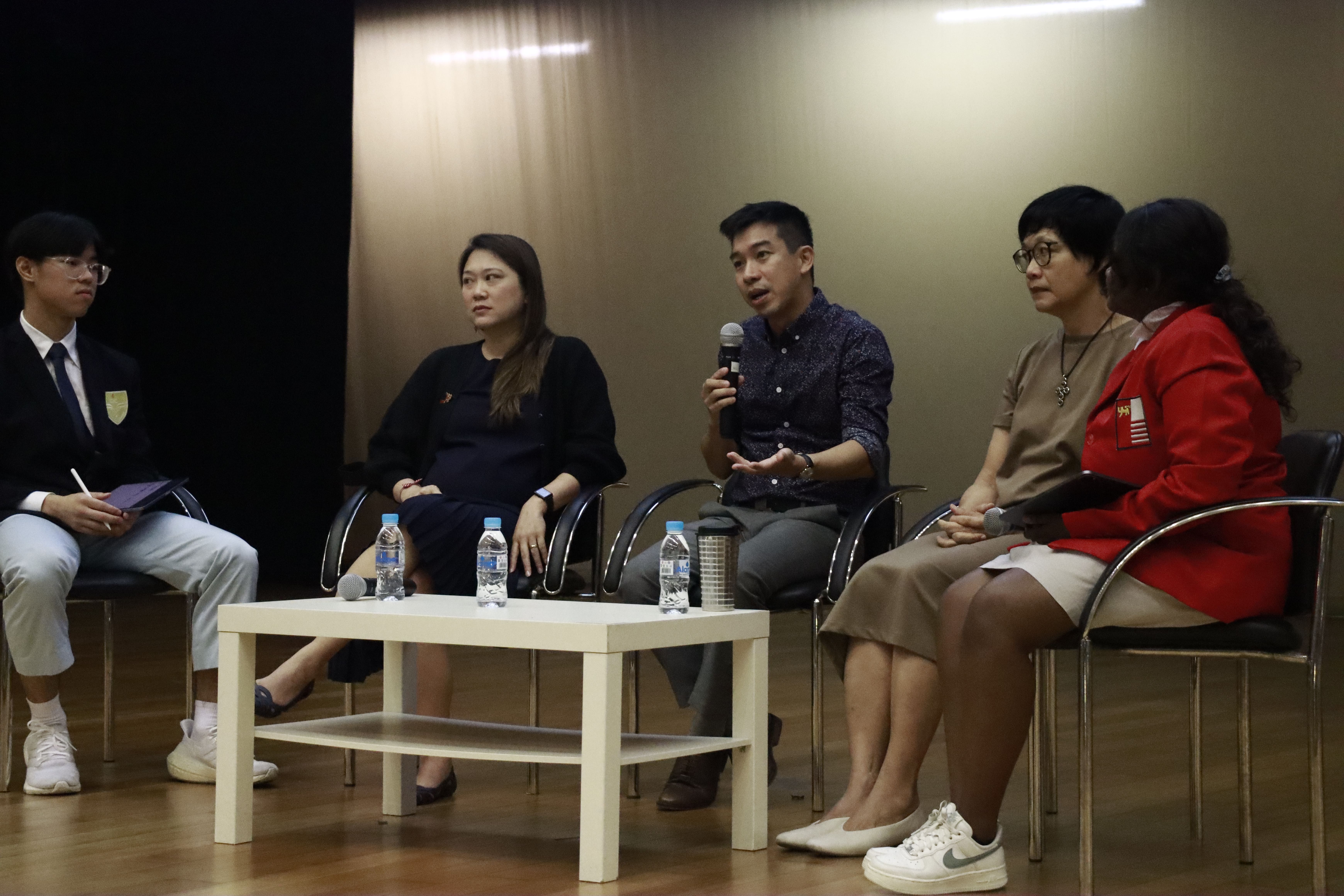 Five people sit in chairs on a stage during a panel discussion, with one man speaking into a microphone.